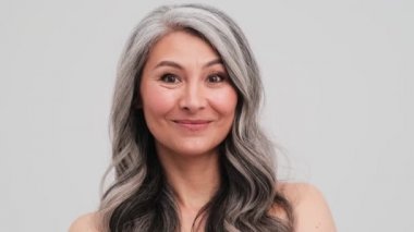 Gülümseyen yaşlı bir kadın, seyirciyi stüdyodaki gri duvarın üzerinde tek başına dikilmeye davet ediyor.