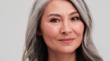 Kameraya gülümseyen yaşlı bir kadının yakın çekim görüntüsü atölyedeki gri duvarın üzerinde izole bir şekilde duruyor.