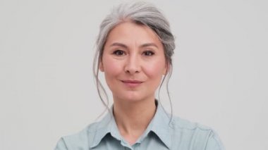 Neşeli yaşlı bir kadının yakın plan görüntüsü ağzını kapatıyor. Stüdyodaki gri duvarın üzerinde tek başına duruyor.