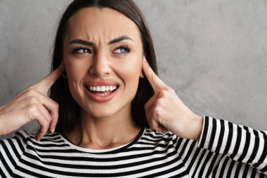 Gri arka plan yüzünden kulaklarını tıkayan gülümseyen genç ve çekici bir kadının yakın çekimi.