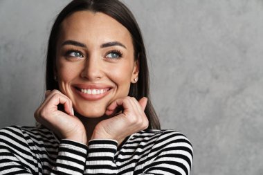 Gri arka planda izole edilmiş uzun saçlı, güzel, gülümseyen esmer bir kadına yaklaş, poz ver, başka tarafa bak.