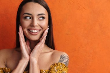 Turuncu arka planda izole edilmiş uzun saçlı, güzel, gülümseyen esmer bir kadına yaklaş, poz ver, yüzünü çevir.