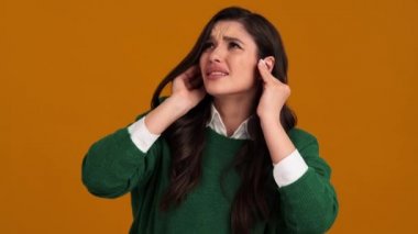 Yeşil kazaklı ve gömlekli kayıp bir kadın turuncu bir stüdyoda dikilirken kulaklarını kapatıyor.