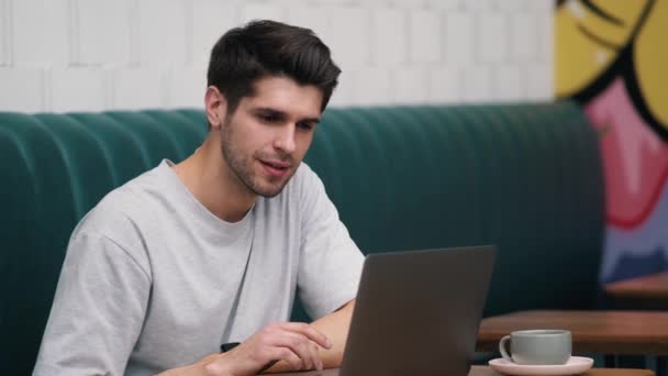Egy Mosolygó Fiatalember Pólóban Nézi Laptop Képernyőjét Miközben Egy Kávézóban — Stock videók