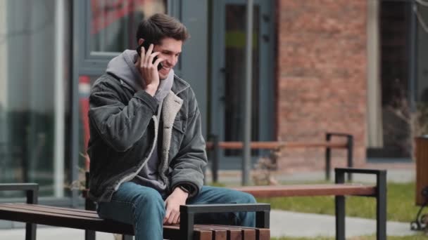 一个穿着连帽衫和斜纹棉布夹克的快乐男人坐在街上的长椅上 一边打电话一边结束通话 — 图库视频影像