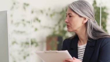 Gri saçlı, evrakları kontrol eden ve ofiste otururken monitöre bakan Asyalı kendine güvenen bir kadının yan görüntüsü.