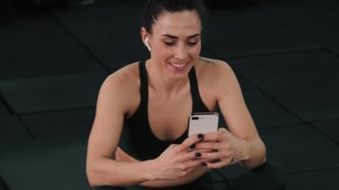 Gülümseyen bir sporcu kadın spor salonundaki yoga minderinde otururken akıllı telefonunu kullanıyor.