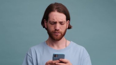 Ciddi bir adam mavi arka planda tek başına duran akıllı telefonunu kullanıyor.