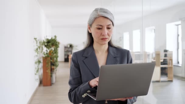 オフィスのラップトップに何かを入力して灰色の髪のスーツに自信のあるアジアの女性 — ストック動画