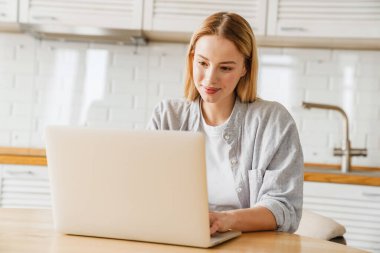 Ev mutfağında otururken gülümseyen ve dizüstü bilgisayar kullanan sarışın kız.