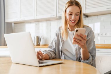 Ev mutfağında otururken bilgisayar ve akıllı telefon kullanan neşeli sarışın kız.