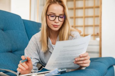 Gözlüklü kız evde kanepede yatarken ödev yapıyor.