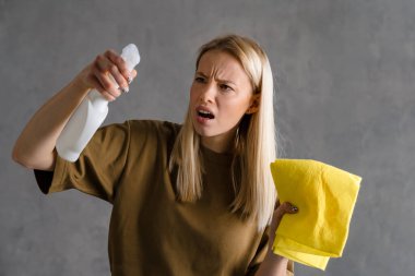 Gri duvarda izole edilmiş temizlik spreyi ve bez parçasıyla poz veren Avrupalı bir kadın
