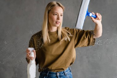 Genç Avrupalı kadın gri duvarda izole edilmiş çizgiler ve spreyle cam temizliyor.