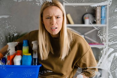 Kafası karışmış sarışın kadın temizlikçi sepet dolusu temizlik malzemesi tutuyor.