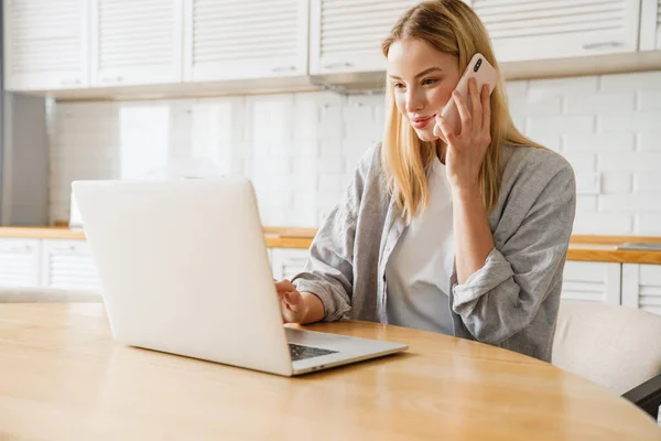 Joyful Blonde Girl Using Laptop Talking Smartphone While Sitting Home — Stock Photo, Image