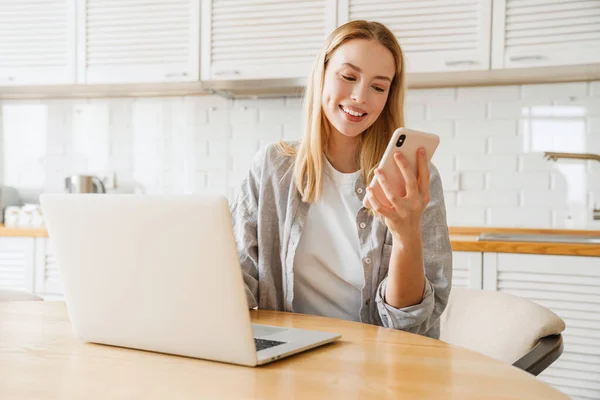 Joyful Blonde Girl Using Laptop Smartphone While Sitting Home Kitchen — Stock Photo, Image