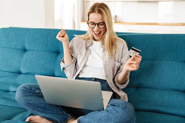 自宅のソファでリラックスしながら ラップトップコンピュータとクレジットカードを介してオンラインで魅力的な若い女性のショッピング笑顔 — ストック写真