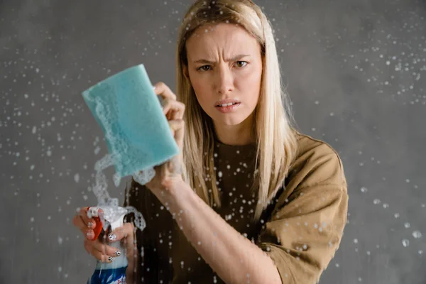 布朗德不高兴的女人用清洁剂和隔着灰墙的海绵洗玻璃 — 图库照片