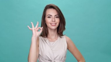 Tişörtlü gülümseyen esmer kadın tamam işareti gösteriyor ve turkuaz arka planda kameraya bakıyor.