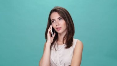 Tişörtlü mutlu esmer kadın turkuaz arka planda akıllı telefonla konuşuyor.