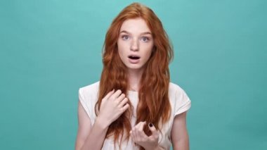 Şaşkın kızıl tişörtlü kadın ağzını kapadı ve turkuaz arka planda kameraya baktı.