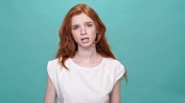 Tişörtlü üzgün kızıl kadın turkuaz arka planı gösterirken bir şey soruyor.