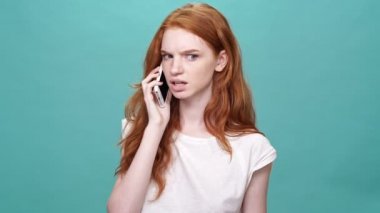 Tişörtlü kızgın kızıl kadın turkuaz arka planda akıllı telefonuyla konuşuyor.