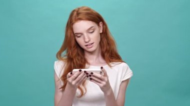 Turkuaz arka planda akıllı telefon kullanan tişörtlü kızıl bir kadın.