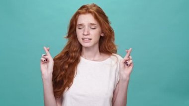 Tişörtlü genç kızıl kadın dua ediyor parmaklarını çapraz yapıp turkuaz arka planda seviniyor.