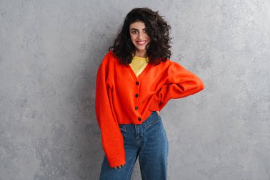 Gri duvarın üzerinden izole edilmiş kameraya poz veren ve gülümseyen güzel esmer kız.