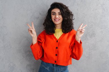 Mutlu güzel kız barış jesti yapıyor ve gri duvarın üzerinden izole edilmiş kameraya gülümsüyor.