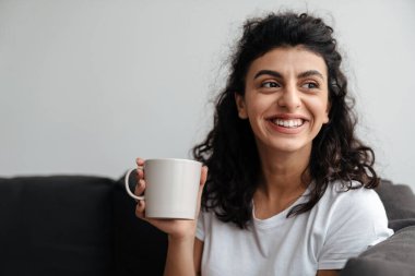 Mutlu esmer kadın evde kanepede otururken gülümsüyor ve kahve içiyor.