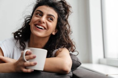 Mutlu esmer kadın evde kanepede otururken gülüyor ve kahve içiyor.