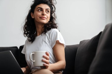 Dizüstü bilgisayarla çalışan ve evde kahve içen mutlu esmer kadın.