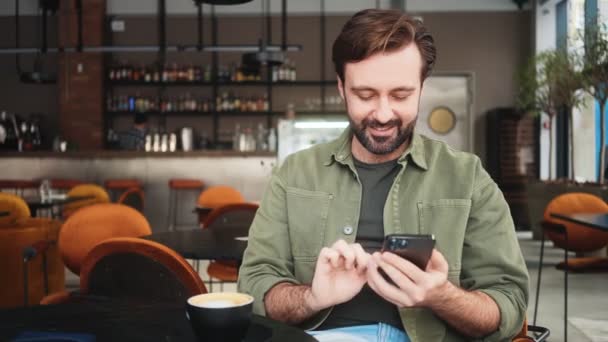 魅力的な男がカフェに座っている彼のスマートフォンを使用しています — ストック動画