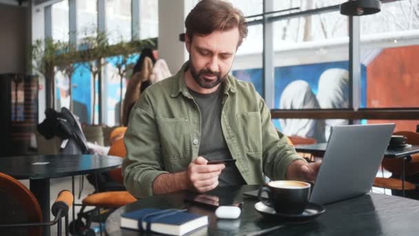 笑顔の男がノートパソコンを使ってるカフェに座ってるクレジットカードを使って — ストック動画