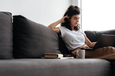 Evde kanepede otururken dizüstü bilgisayarla çalışan odaklanmış genç bir kadın.