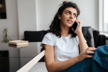Odaklanmış esmer kadın evde otururken cep telefonuyla konuşuyor.