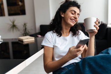 Kahkaha atan esmer kadın evde kahve içerken cep telefonu kullanıyor.