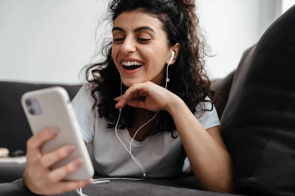 一个快乐的黑发女人躺在沙发上 用手机在耳机里 — 图库照片