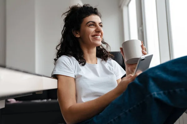 自宅でコーヒーを飲みながら携帯電話を使ってブルネットの女性を笑って — ストック写真