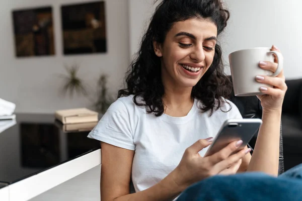 Riendo Morena Mujer Usando Teléfono Móvil Mientras Bebe Café Casa — Foto de Stock