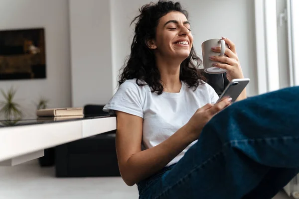 自宅でコーヒーを飲みながら携帯電話を使ってブルネットの女性を笑って — ストック写真