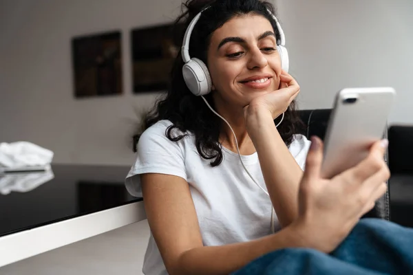 坐在家里用手机在耳机里快乐的黑发女人 — 图库照片