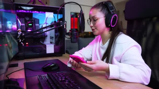眼鏡とヘッドフォンを身に着けている魅力的な女の子のゲーマーは 自宅に座って彼女のスマートフォンを使用している間 プロの強力なコンピュータ上でオンラインビデオゲームをストリーミングしています — ストック動画