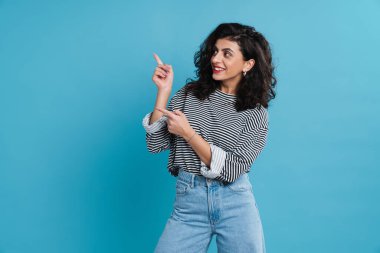 Neşeli güzel kız gülümsüyor ve mavi arka planda izole edilmiş parmakları işaret ediyor.