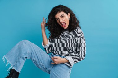 Heyecanlı kız mavi arka planda dans ederken ve çığlık atarken boynuz hareketi yapıyor.