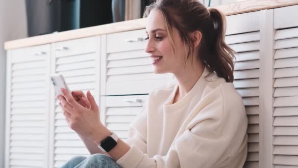 Μια Ευχαριστημένη Ξανθιά Γυναίκα Χρησιμοποιεί Smartphone Της Καθισμένη Στο Σπίτι — Αρχείο Βίντεο