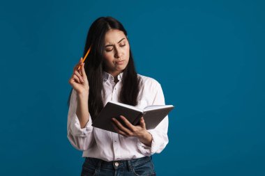 Düşüncelere dalmış güzel Asyalı kız mavi arka planda izole edilmiş notlar yazıyor.